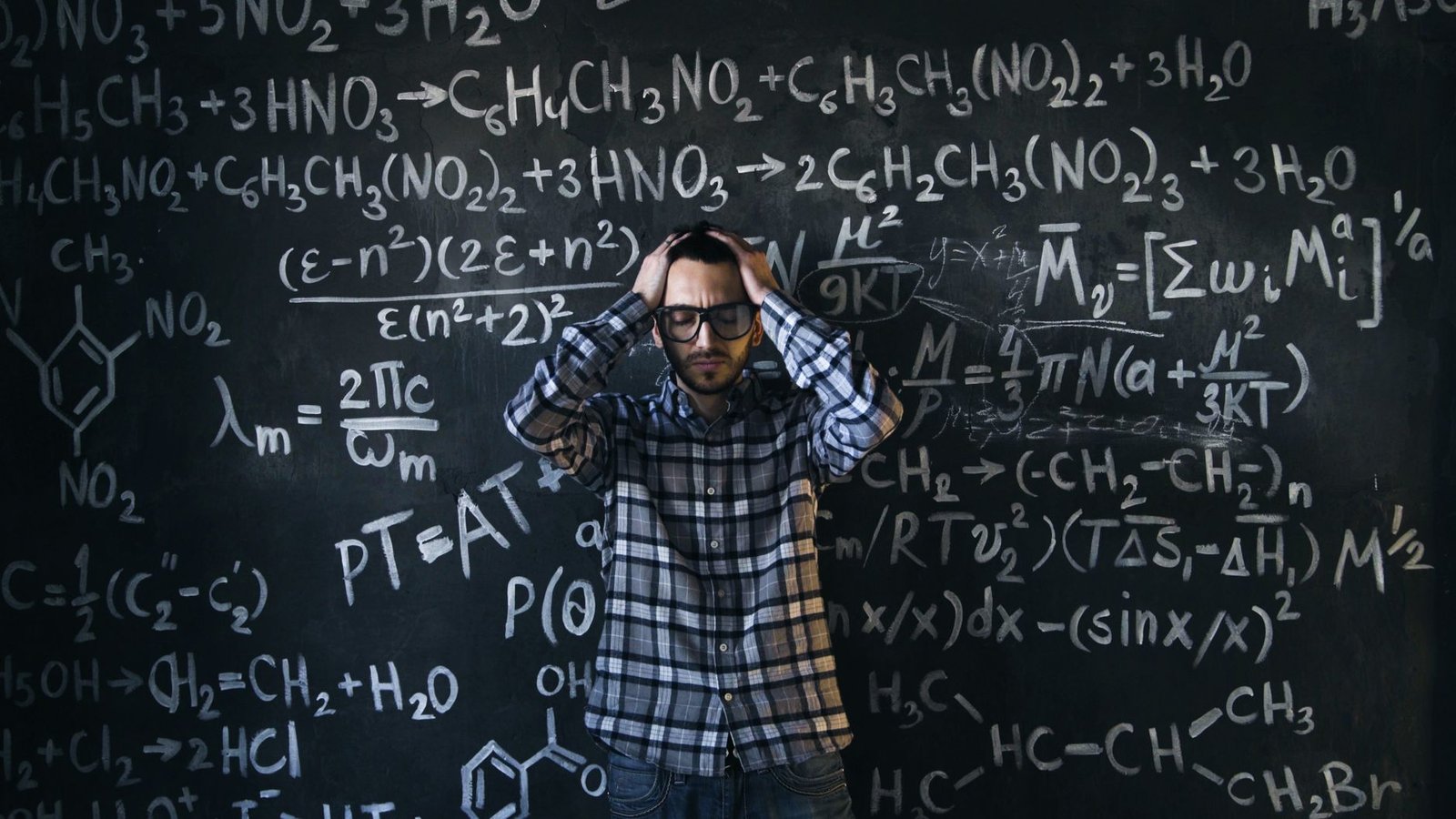 man in eyeglasses holding his head next to blackboard with math equations