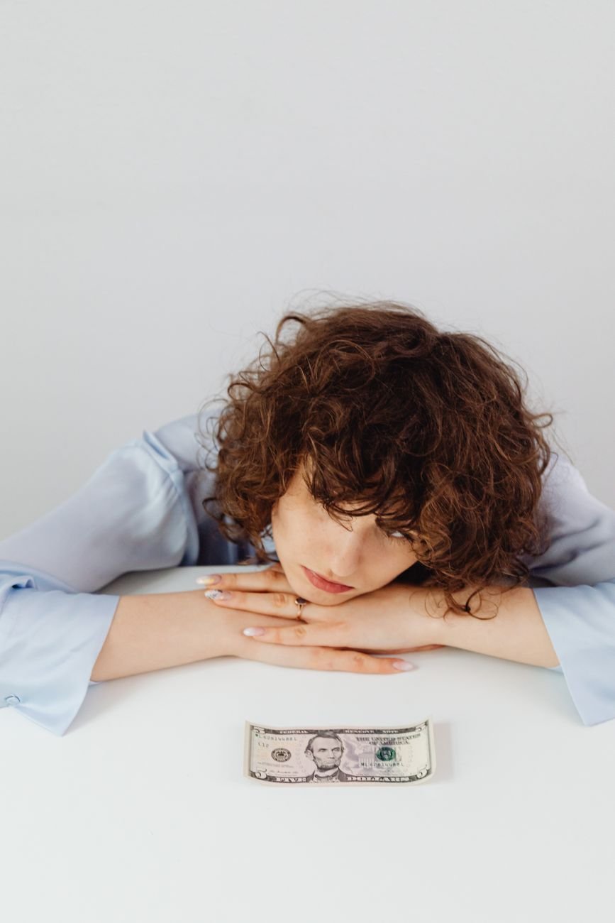 a woman in blue long sleeves resting her head on the table with paper money while looking afar