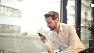 photo of man in white dress shirt holding phone near window