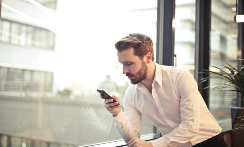 photo of man in white dress shirt holding phone near window